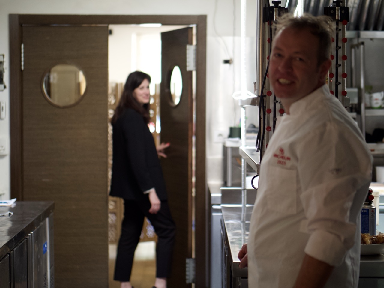 José Andrés en primer plano en la cocina y Jessica pasando hacia la sala de La Capilleta